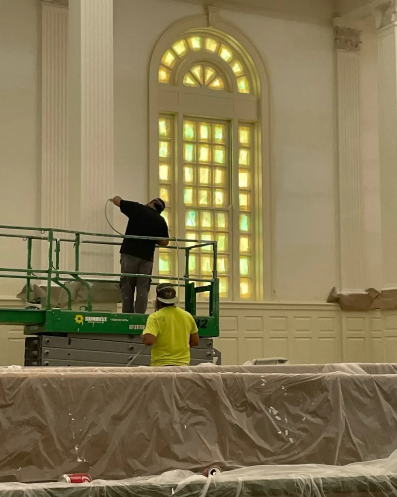 Two painters on a powered lift painting tall arched windows in a church