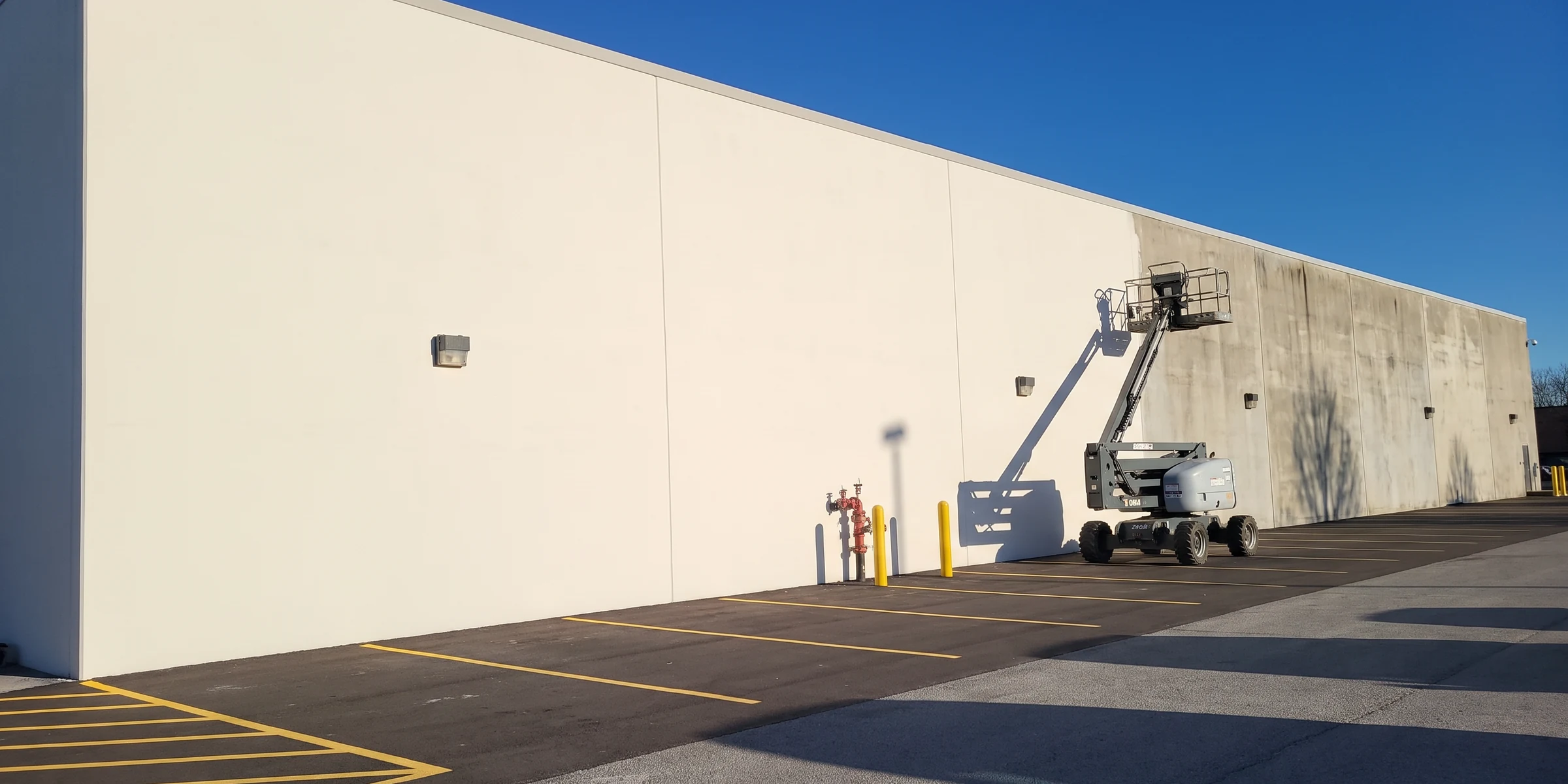 A warehouse wall half recoated in clean white, the far end still the original weathered gray, with a boom lift at the line
