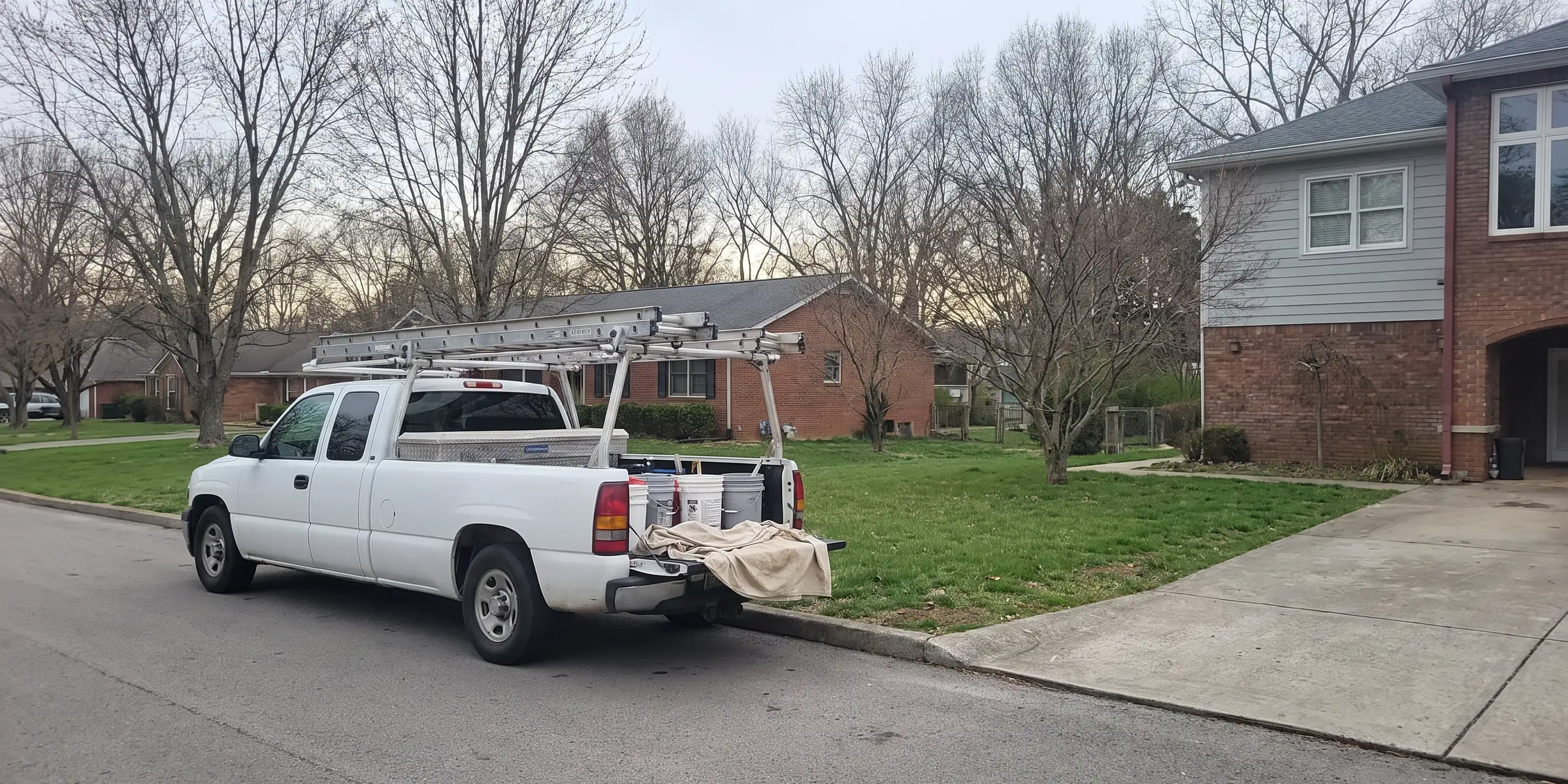 A white work pickup parked at the curb outside a house, extension ladders on the rack and buckets in the bed at the end of the day