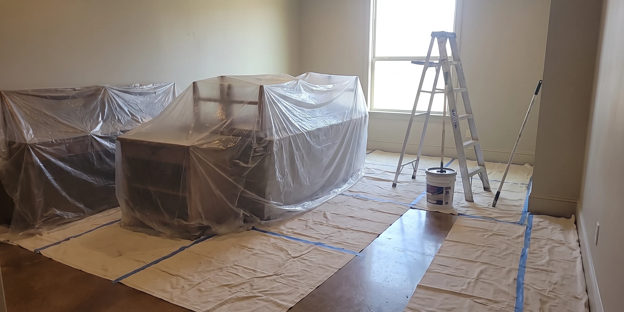 A room prepared before any paint goes on, furniture wrapped in plastic and drop cloths taped down over the floor